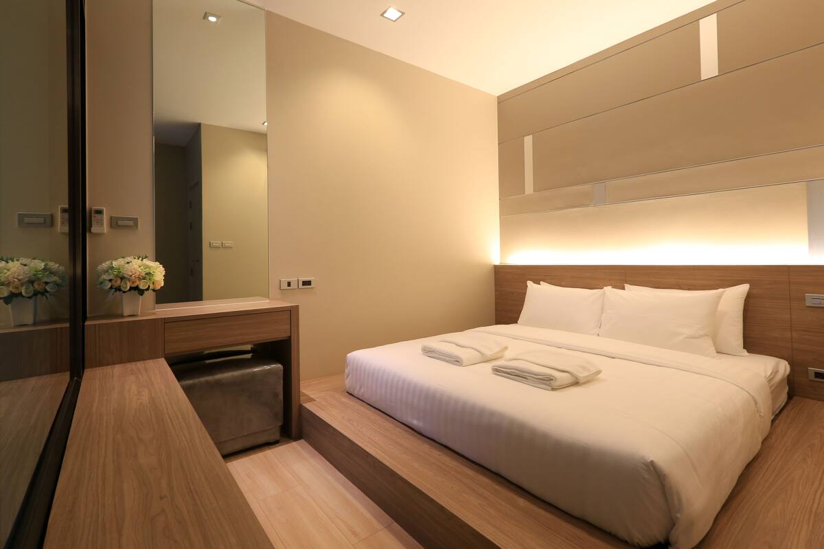 Una habitación de hotel moderna con una cama grande, una mesita de noche de madera, un espejo y una pequeña zona de baño.