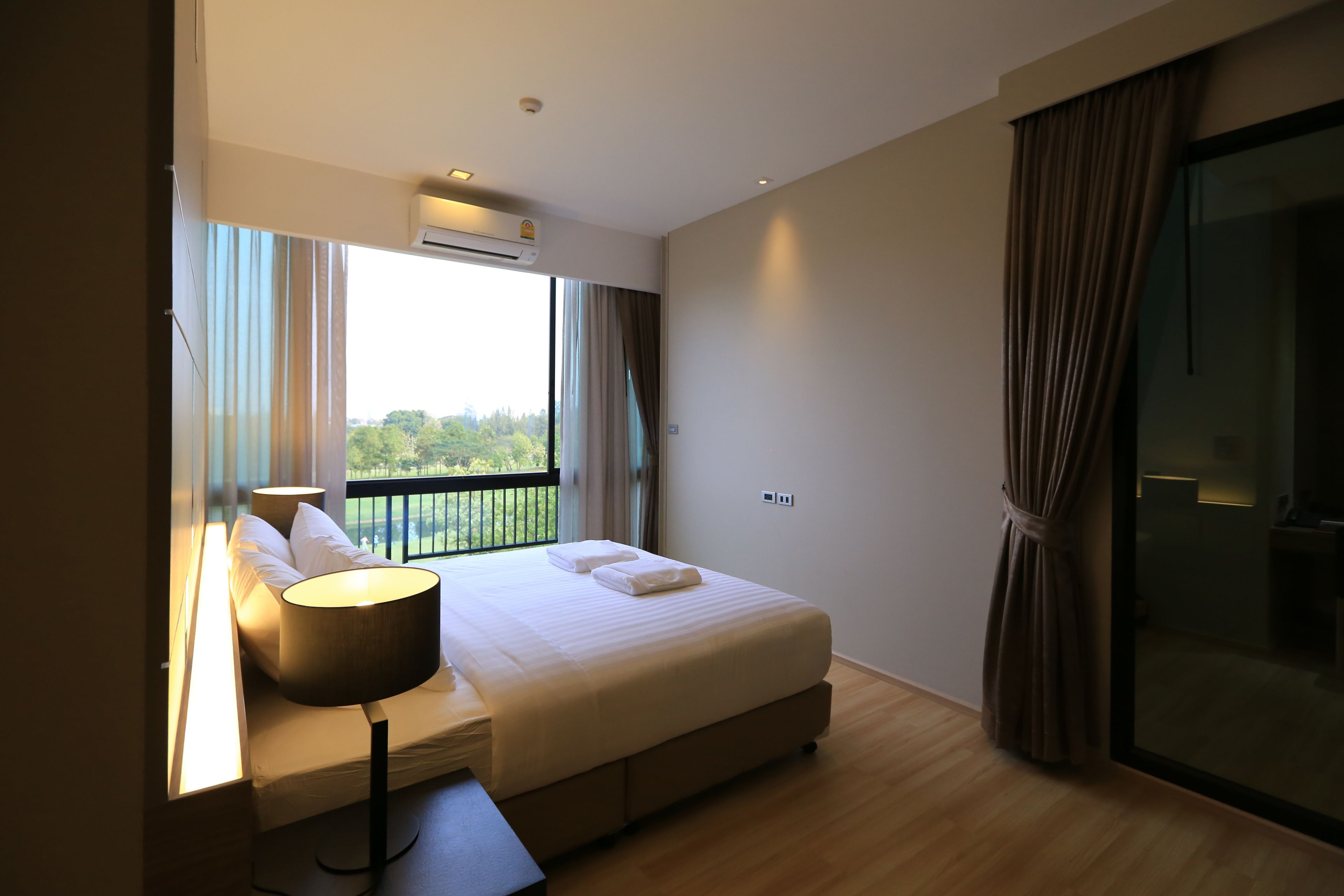 A modern hotel room with a large bed, a bedside table with a lamp, a window with curtains, and a balcony view.