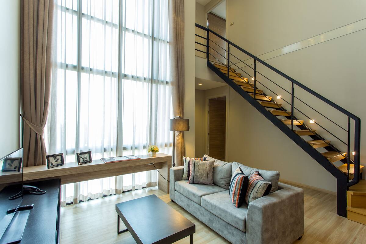Una sala de estar moderna con un sofá, una mesa de centro y un escritorio con una computadora y fotografías. Una escalera con barandilla conduce a un nivel superior.