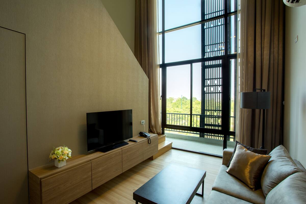 Una sala de estar moderna con un televisor de pantalla plana sobre un mueble de madera, un sofá con cojines, una mesa de centro y un amplio ventanal con vistas a zonas verdes.