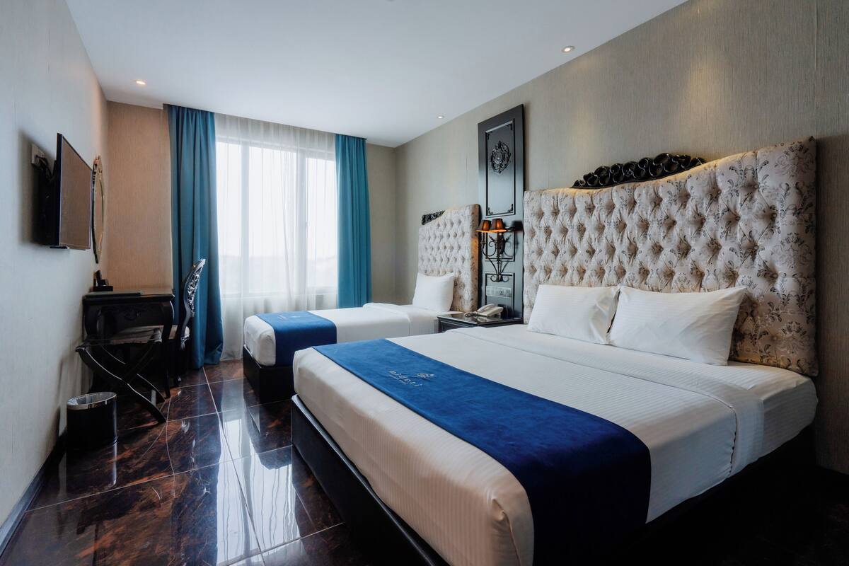 A hotel room with two beds, a desk, a chair, a TV, and a large window with curtains.