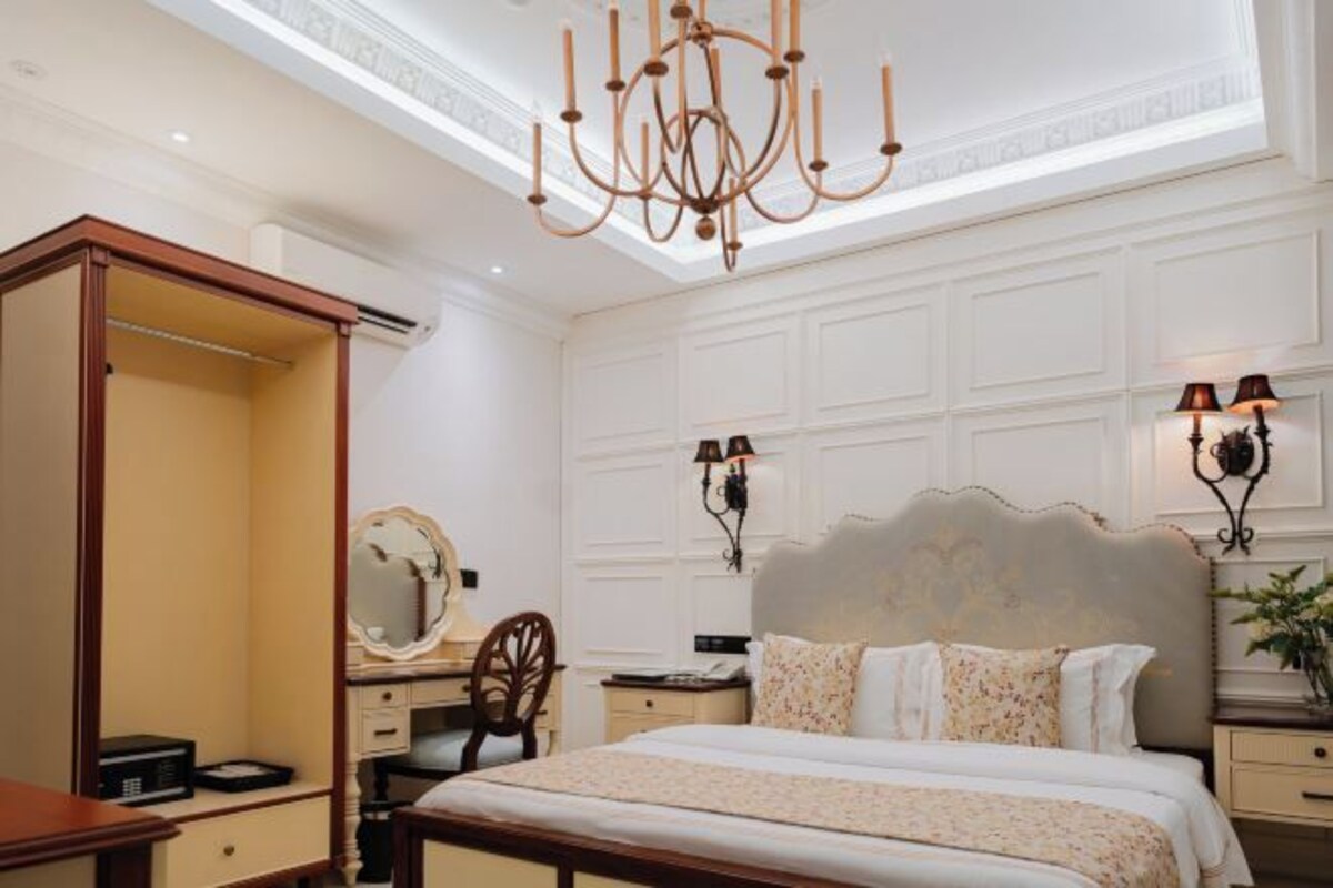 A bedroom with a large bed, a vanity with a mirror, a wooden wardrobe, and a chandelier.