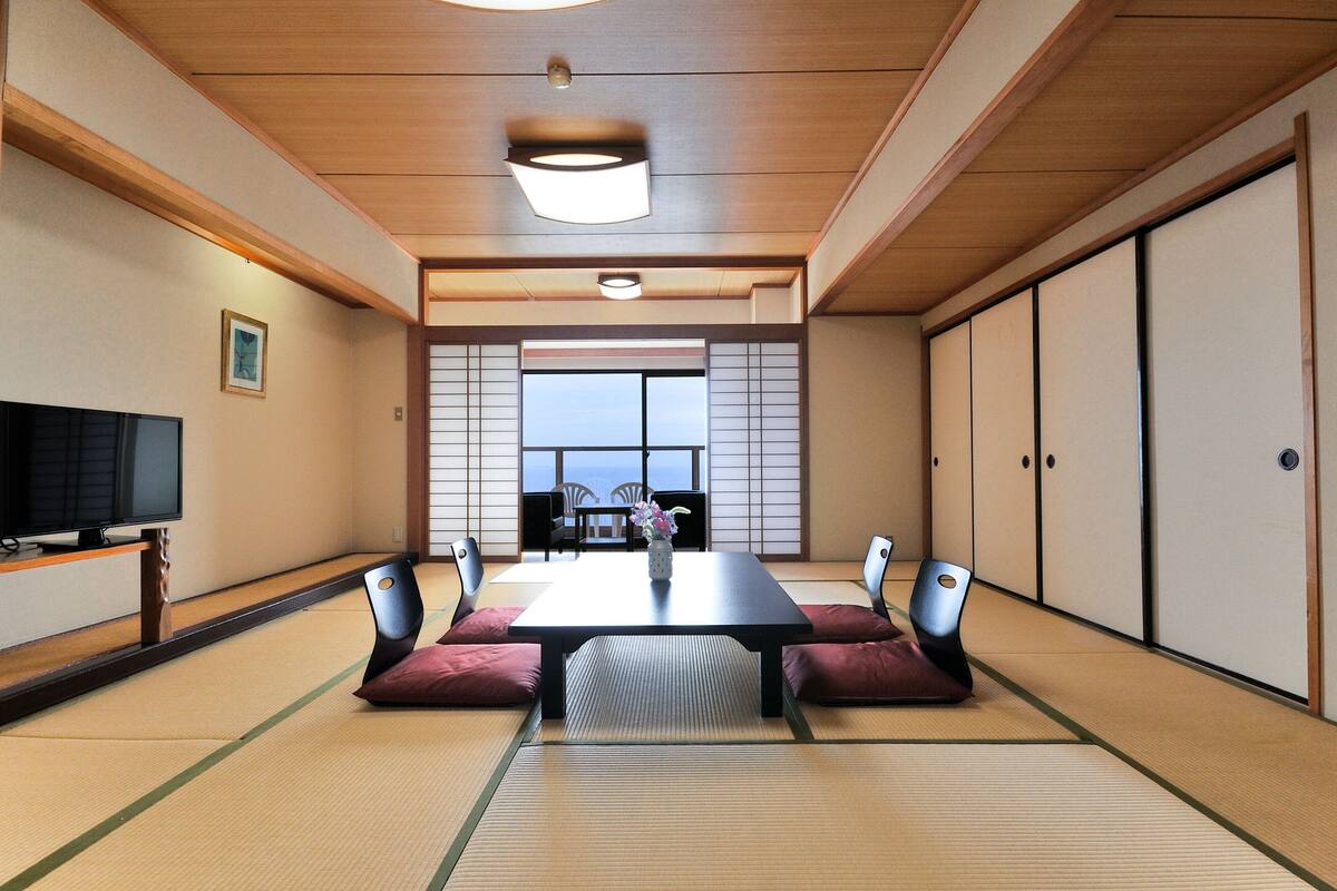 A traditional Japanese room with tatami flooring, a low table, cushions, sliding doors, and a television.