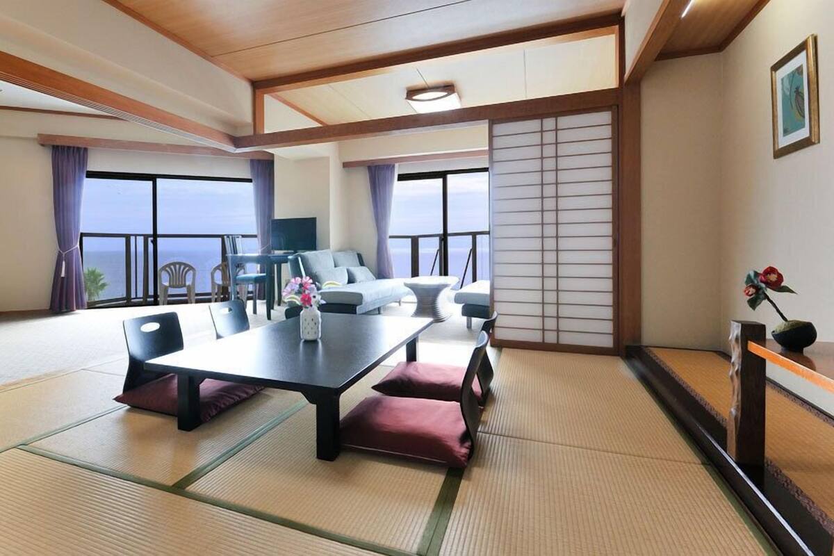 A hotel room with tatami mats, a sofa, a dining table, and a view of the sea.
