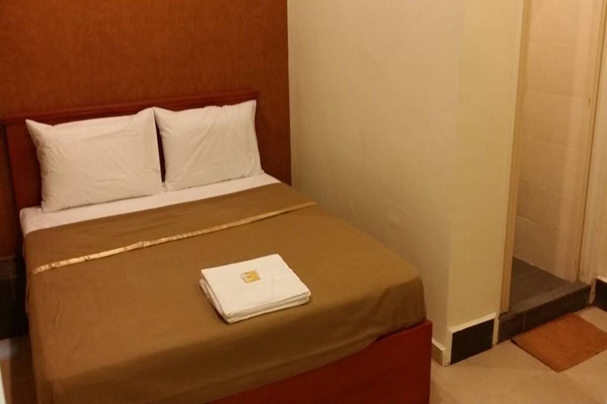 Cama bem arrumada com travesseiros brancos e colcha marrom, uma toalha branca dobrada sobre a cama e cabeceira de madeira.