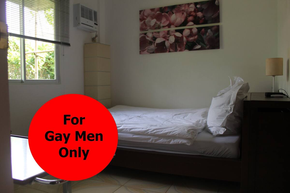 Une chambre individuelle avec un lit simple, une fenêtre, un climatiseur, une lampe fixée au mur et un tableau représentant des fleurs.