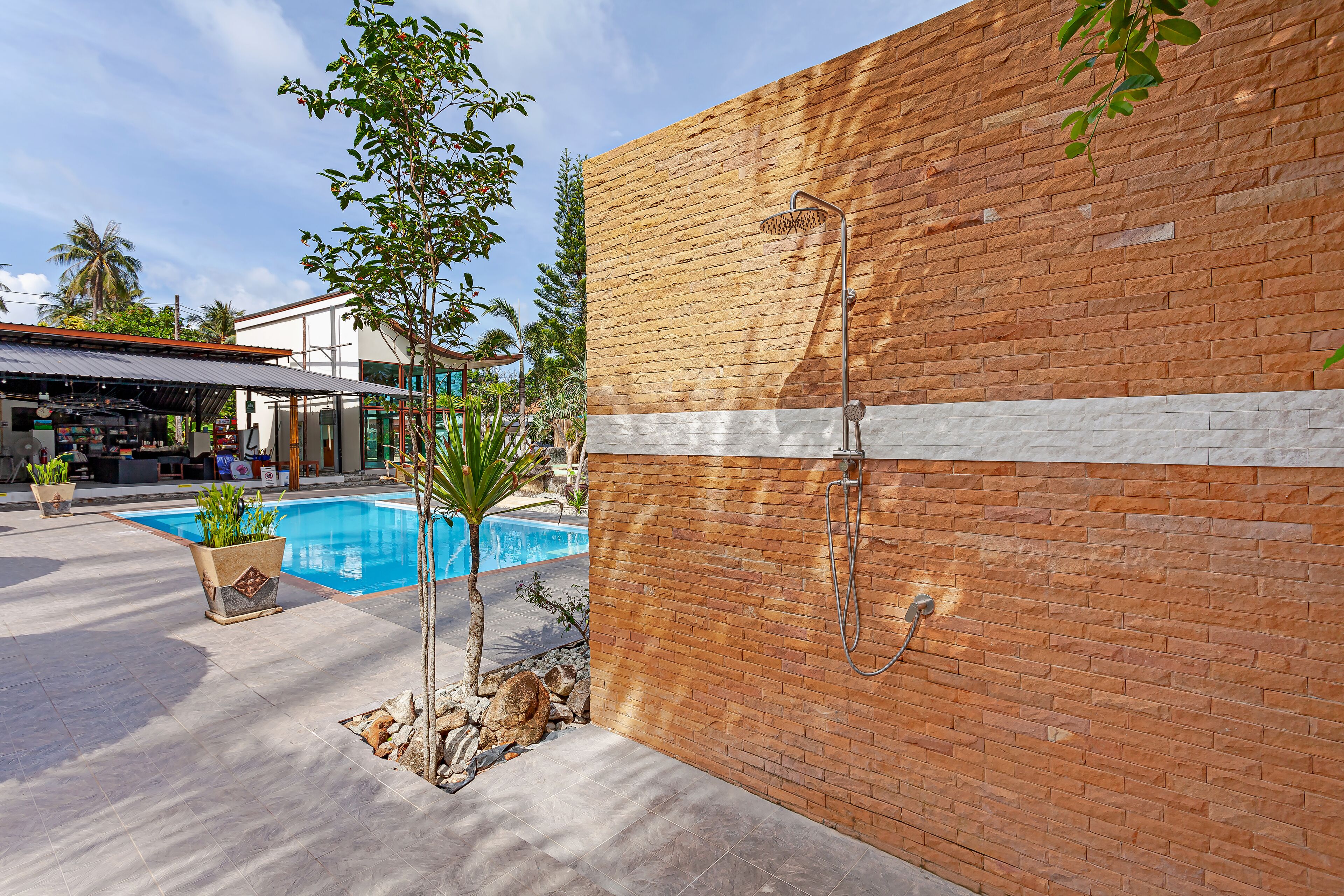 Zona de piscina con pared de ladrillo, ducha metálica y plantas en macetas.