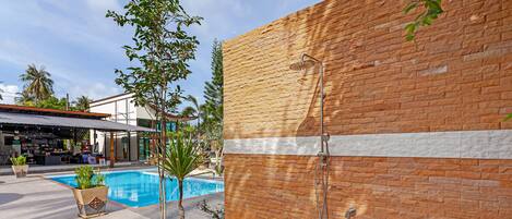Zona de piscina con pared de ladrillo, ducha metálica y plantas en macetas.