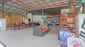 Un espacio al aire libre techado con mesas y sillas, una estantería llena de libros infantiles y una variedad de objetos en repisas.