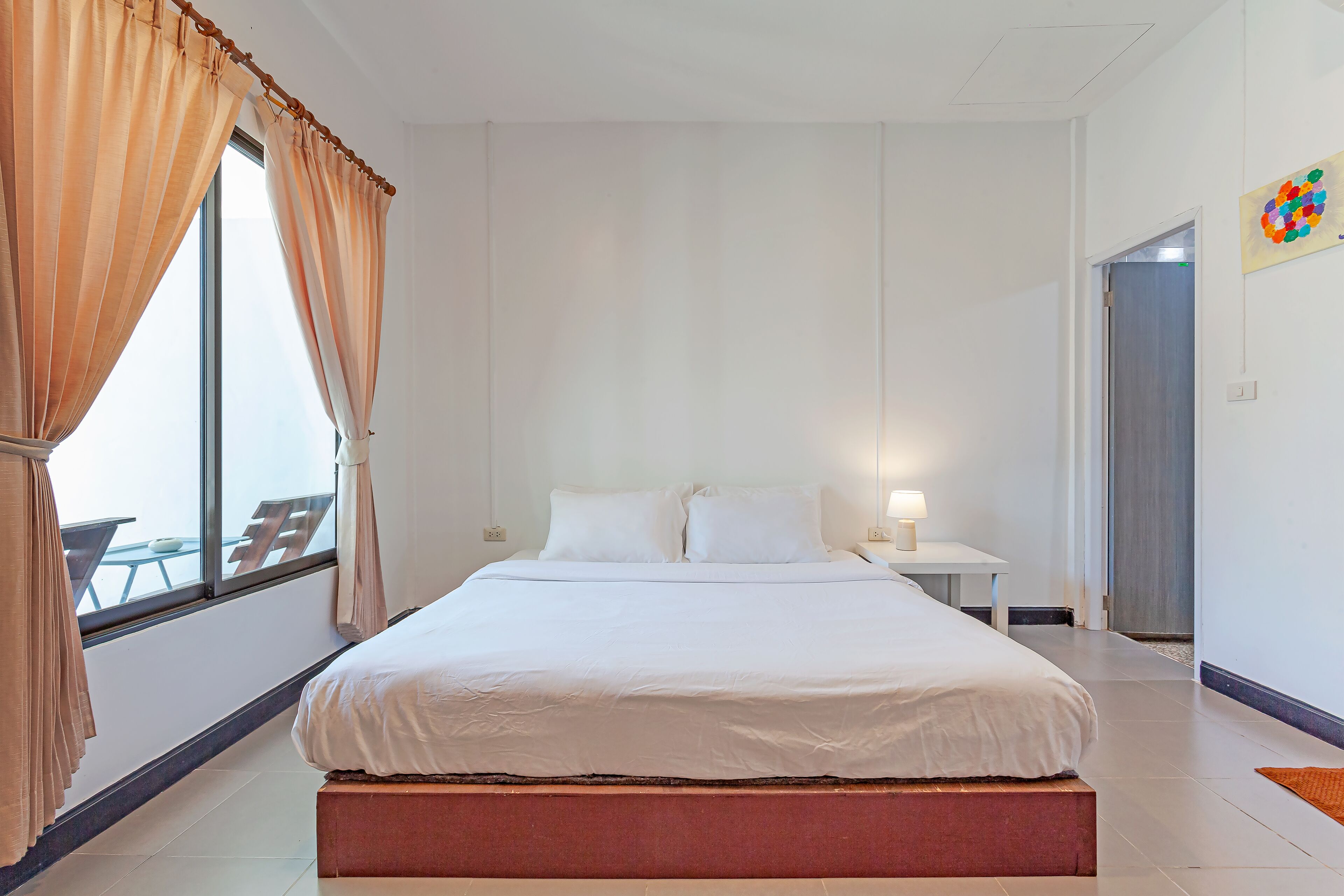 Habitación de hotel con una cama grande, una mesita de noche con lámpara y vistas al exterior a través de una ventana con cortinas.