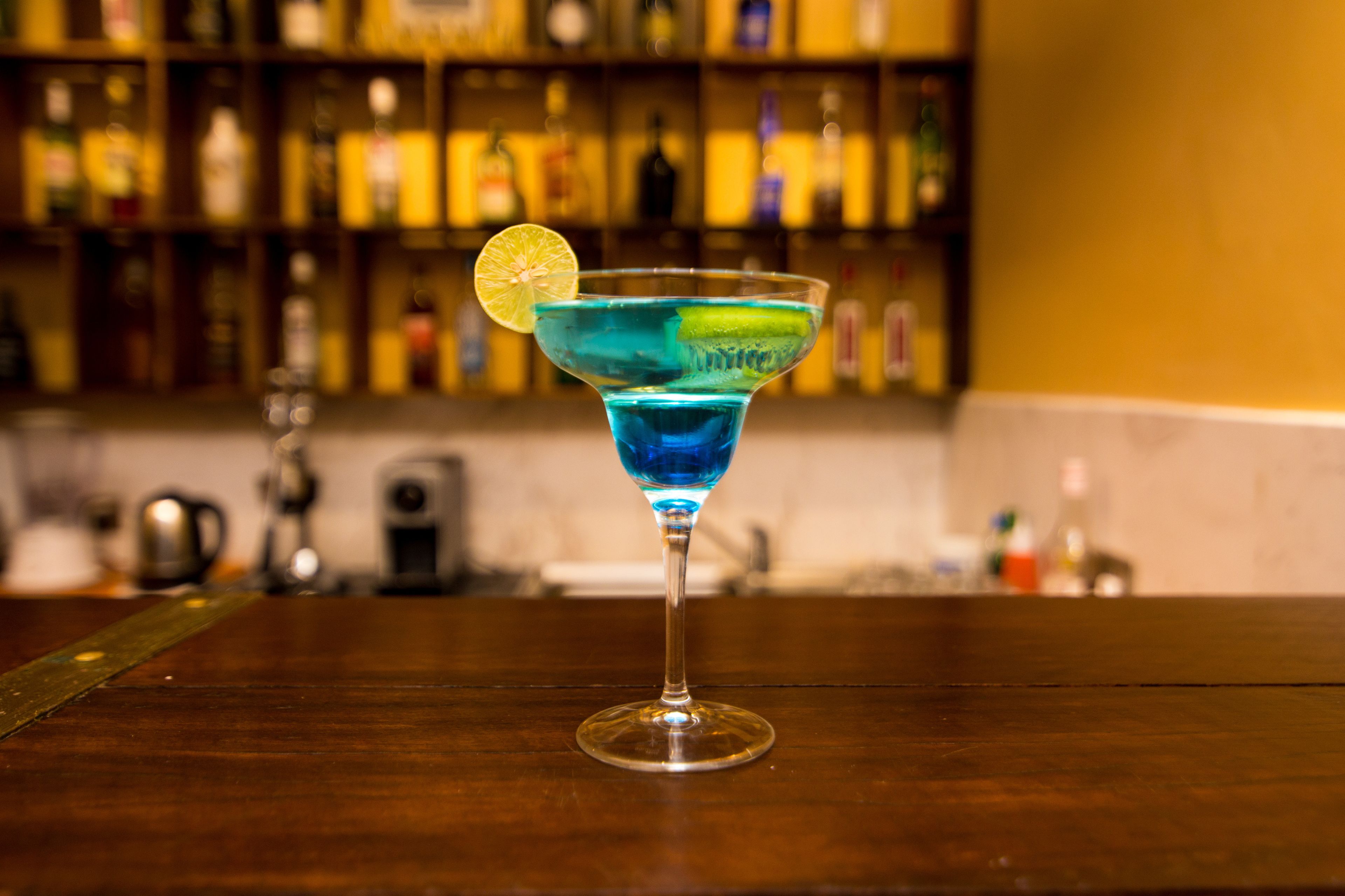Un cocktail bleu servi dans un verre à martini, agrémenté d’une tranche de citron, sur le comptoir en bois d’un bar.