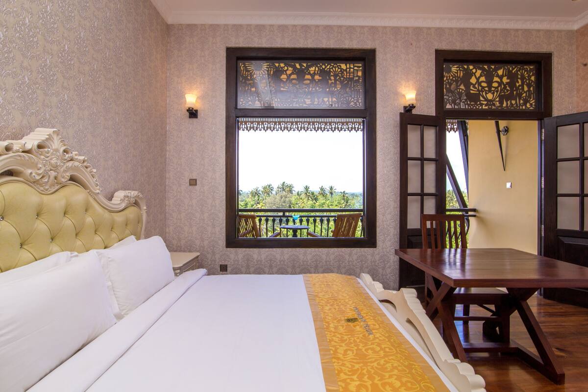 A hotel room with a large bed, a wooden table, and a balcony with a view of greenery.