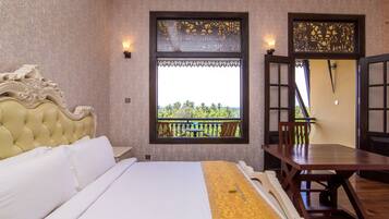 A hotel room with a large bed, a wooden table, and a balcony with a view of greenery.