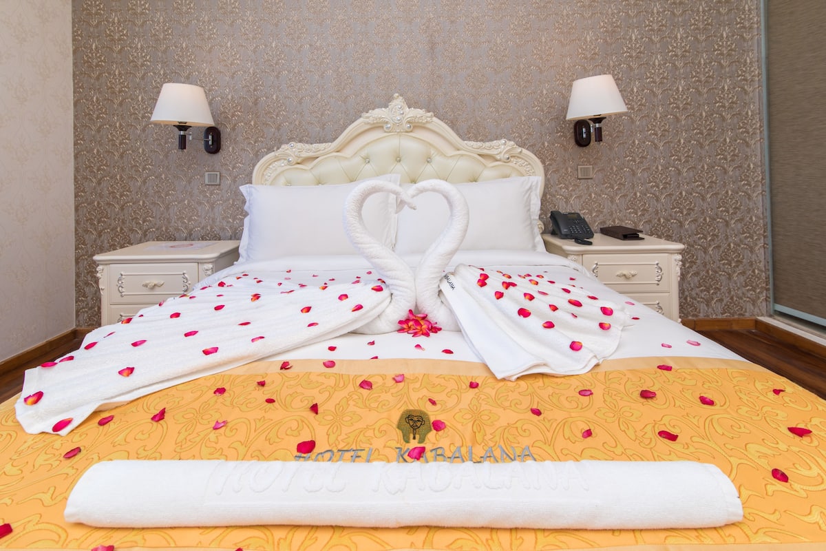 A hotel room with a bed featuring a heart-shaped towel arrangement, a decorative headboard, and a floral-patterned bedspread.