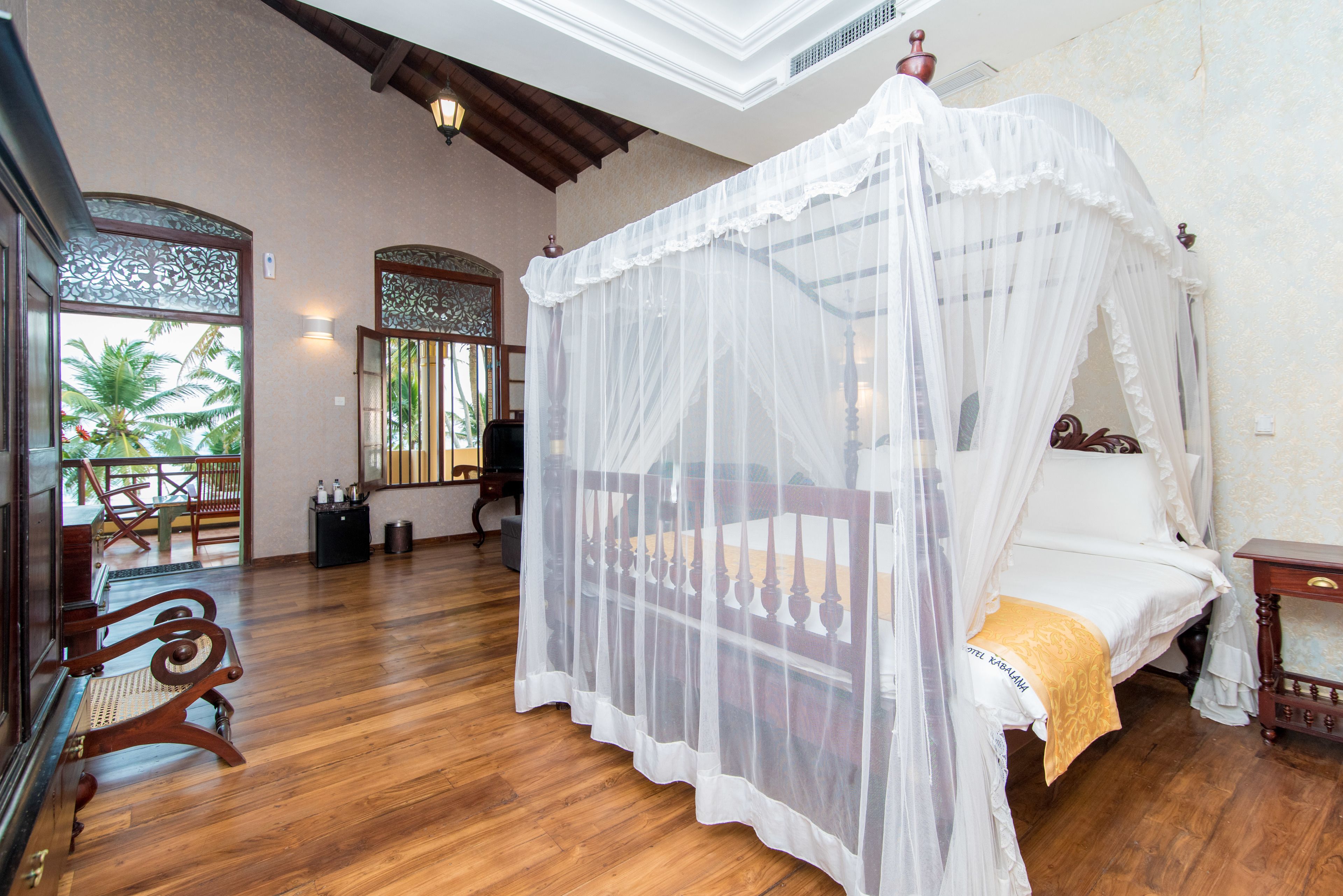 A room with a four-poster bed, wooden flooring, and a balcony with a view of palm trees.