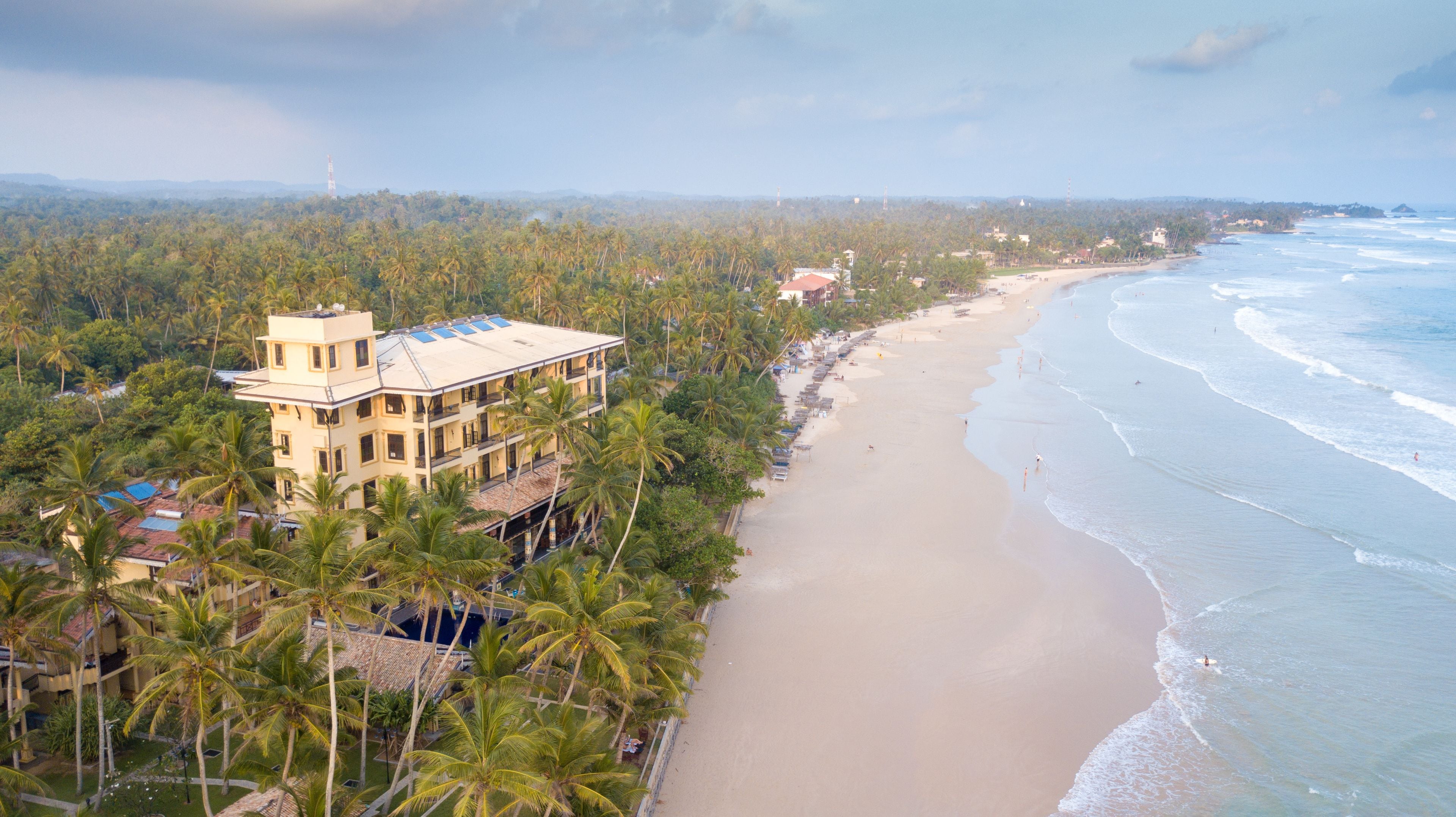 A beachfront hotel with multiple floors, surrounded by palm trees and a sandy beach.