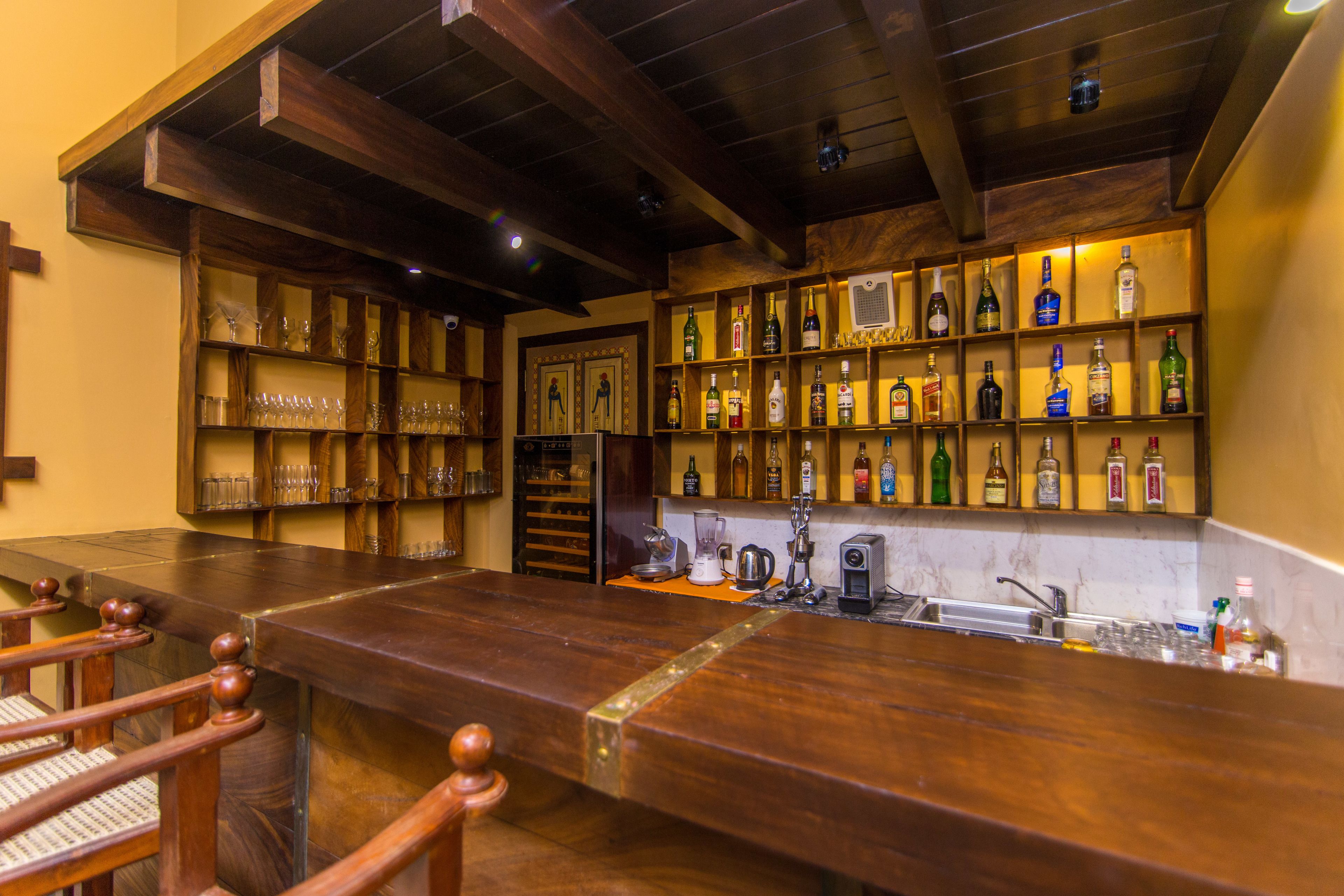 A bar area with wooden shelves displaying various bottles and glasses, a long counter, and a wooden ceiling.