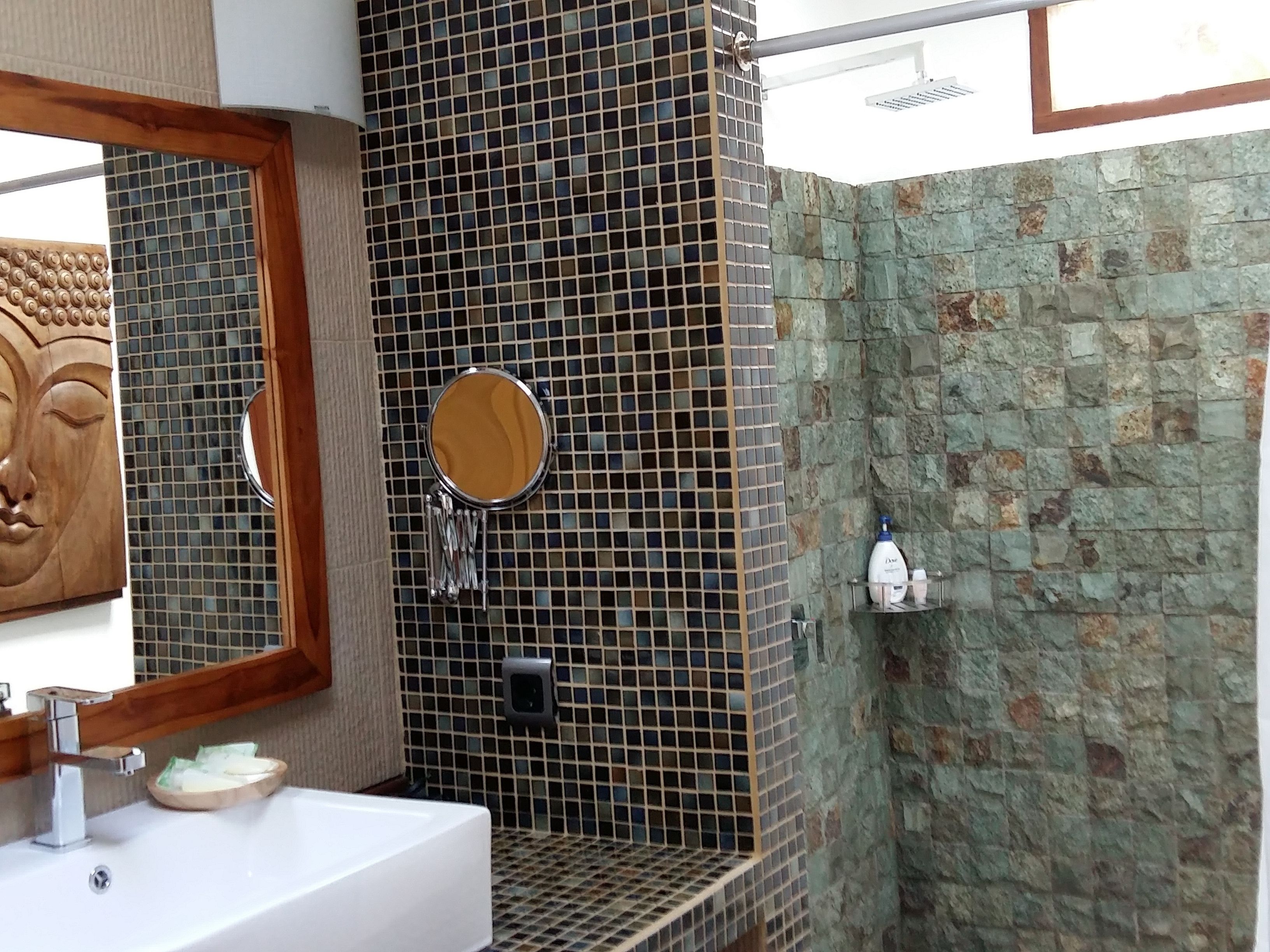 A bathroom with a white sink, a wooden-framed mirror, a round mirror, and a shower area with a stone wall.