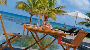 A wooden table with a basket of bread, a glass of orange juice, and a bottle of water by a poolside with palm trees and a view of the ocean. - White Oaks Villas (Pointe Aux Piments)