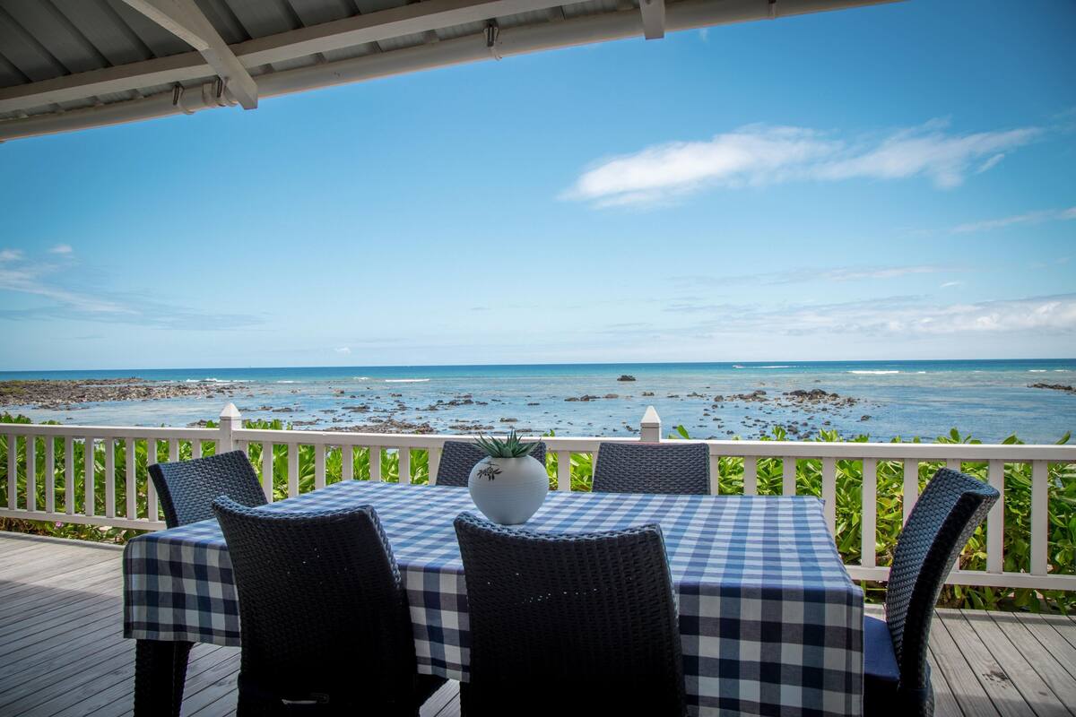 A covered outdoor seating area with a checkered table and chairs, overlooking a rocky beach and the ocean.