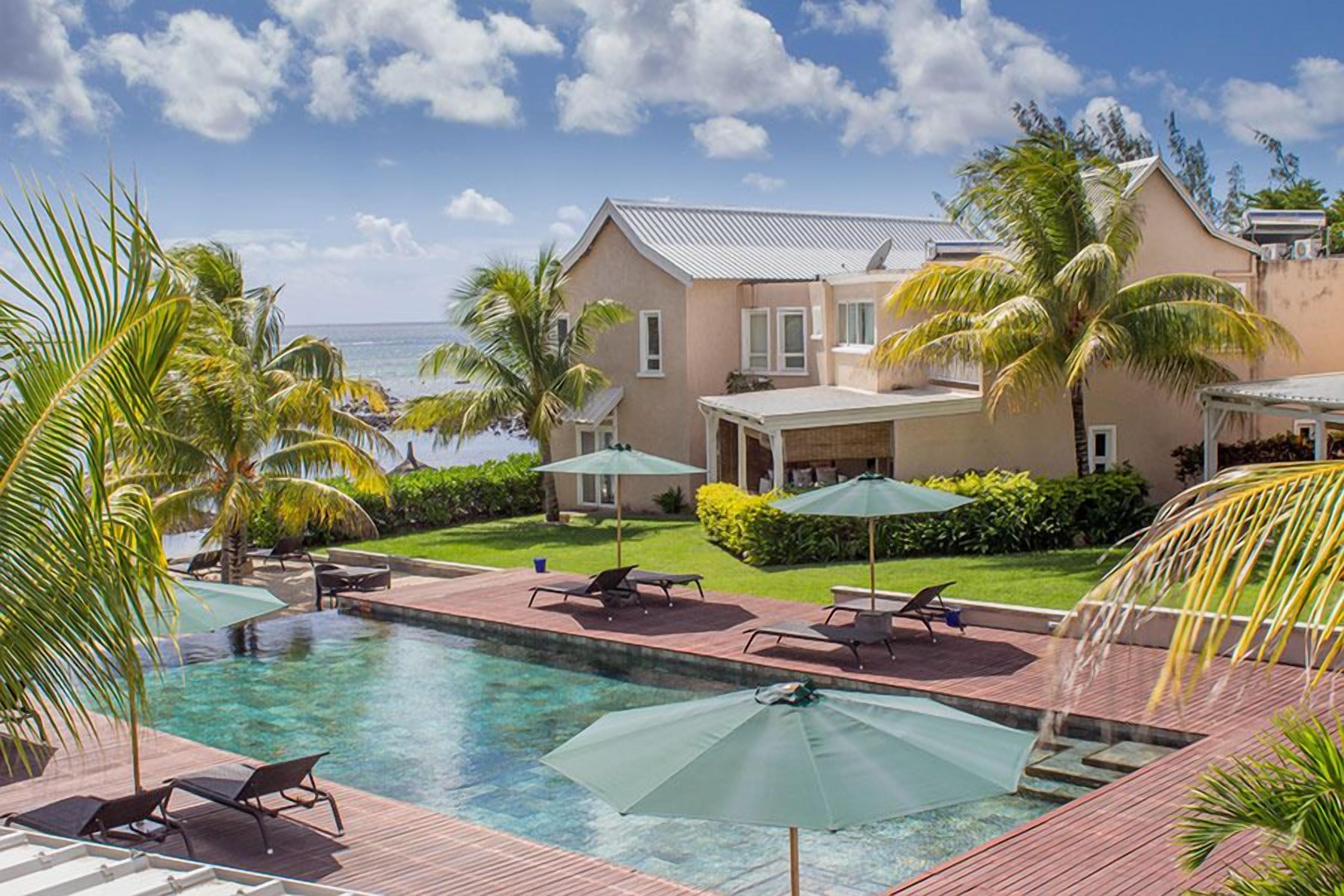Un espace piscine avec des chaises longues et des parasols, une maison dotée d’un patio couvert et des palmiers.