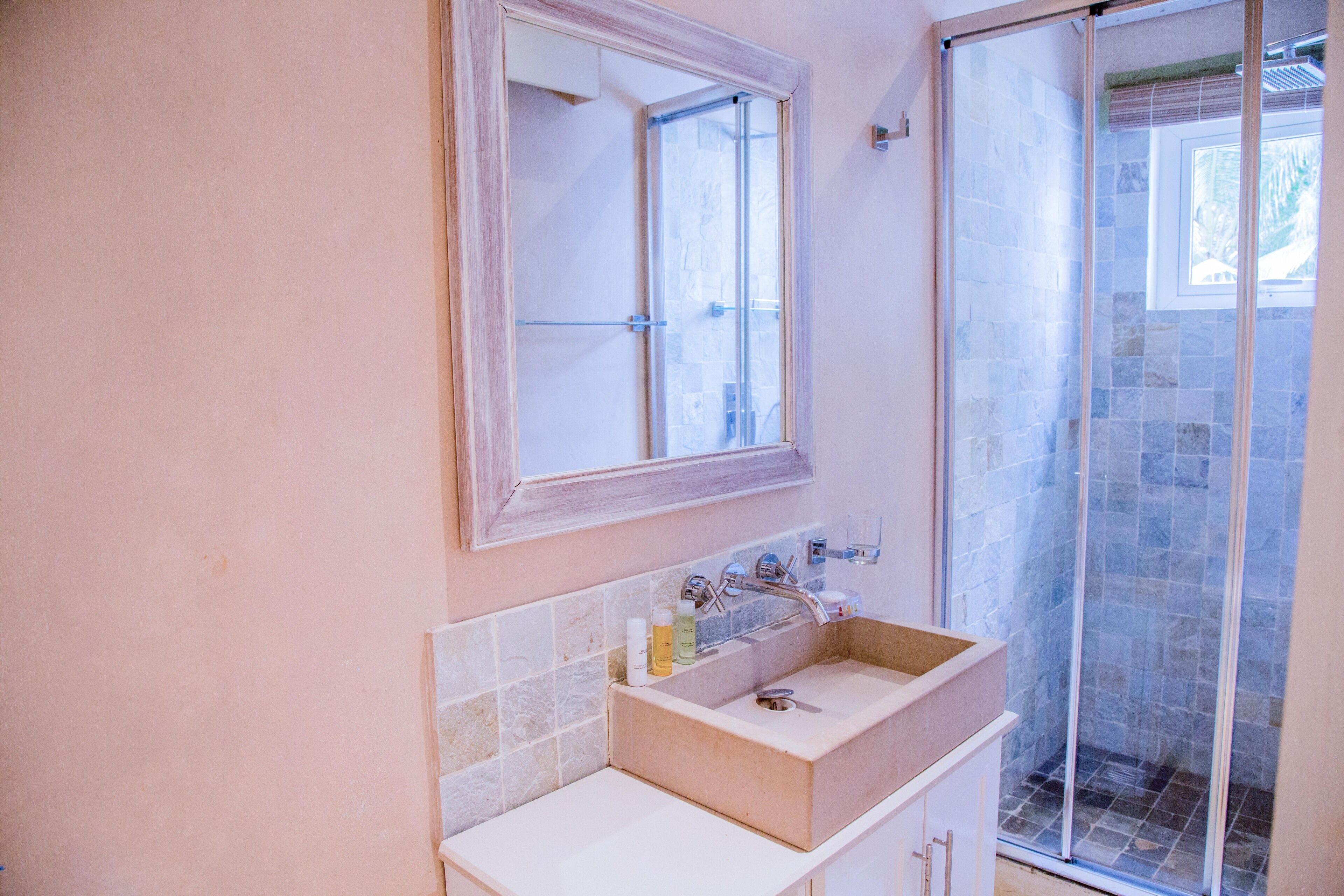 Un baño con lavamanos color crema, un espejo encima y una zona de ducha con azulejos de piedra.