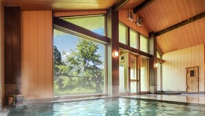 A spacious indoor swimming pool with clear glass walls, wooden beams, and a view of trees outside.