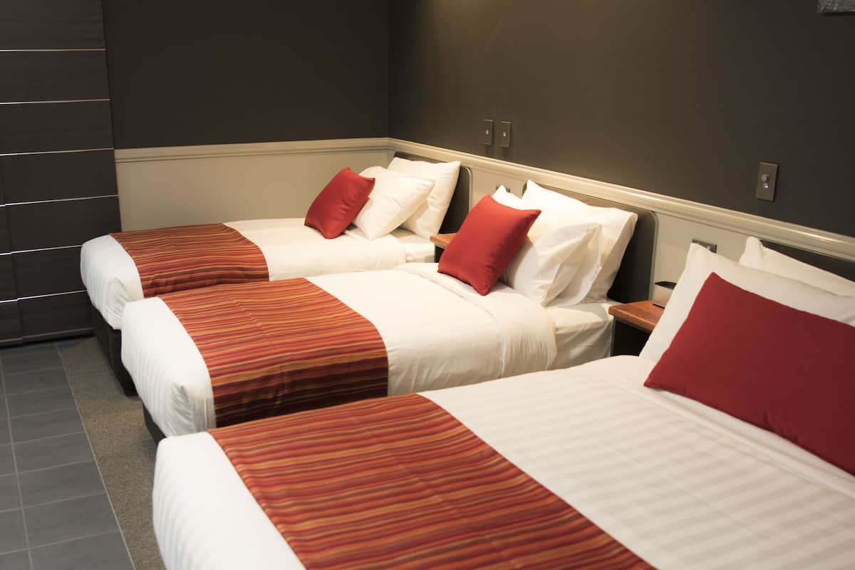 Une chambre d’hôtel avec trois lits, chacun recouvert d’une literie rouge et blanche, et un mur de couleur sombre.