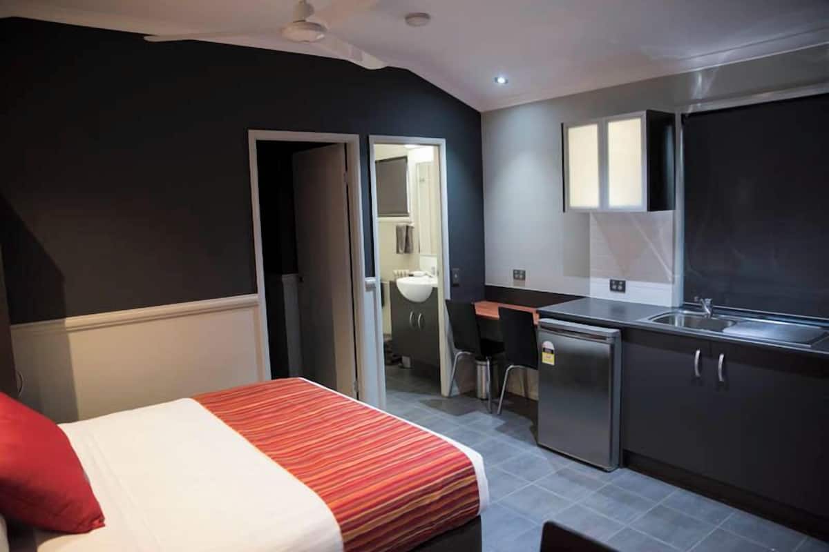 Une chambre d’hôtel de taille réduite comprenant un lit, une petite kitchenette, une salle de bain et une porte donnant sur une autre pièce.