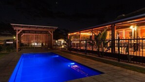 A swimming pool with a wooden deck area, a covered seating area with glass walls, and a fenced patio. - The Rocks Motel (Charleville)