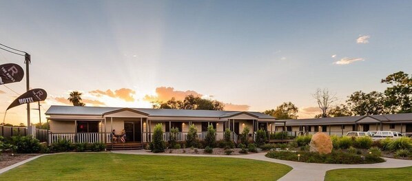 A motel with a covered outdoor seating area, a well-maintained lawn, and a clear sky.