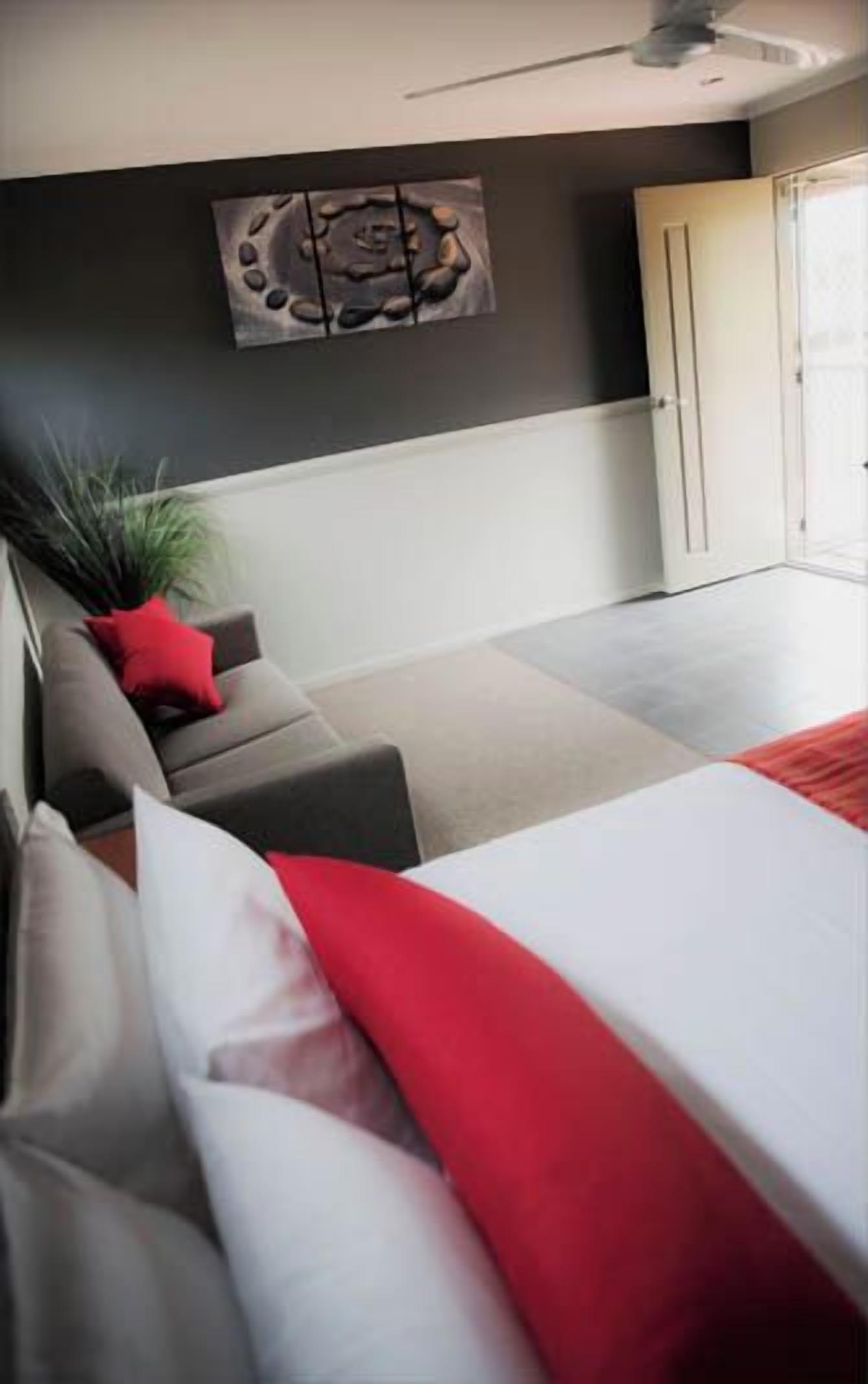A modern bedroom with a bed, a grey sofa, a red pillow, a potted plant, and a door leading to a balcony.