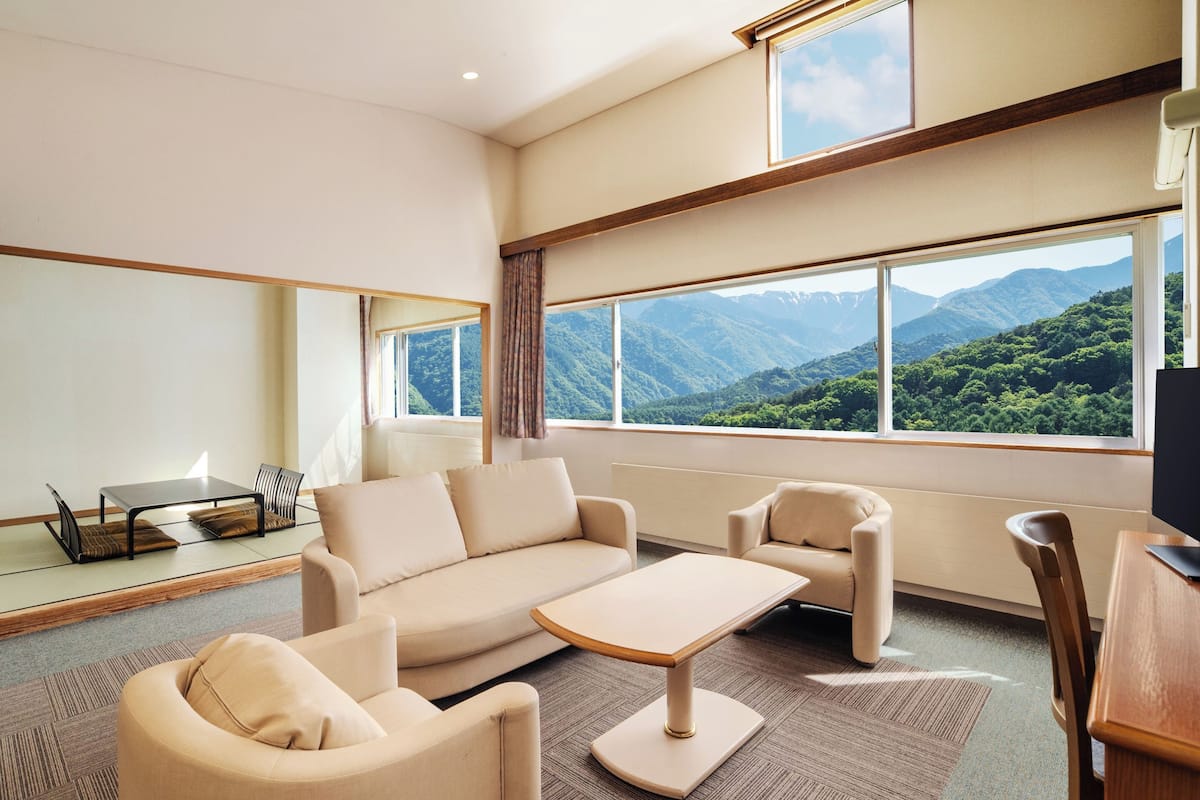 A modern hotel room with a sofa, chairs, a small table, and large windows offering a mountain view.