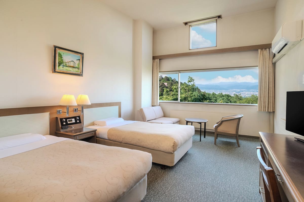 A hotel room with two beds, a desk, a chair, a TV, and a window with a view of greenery and mountains.