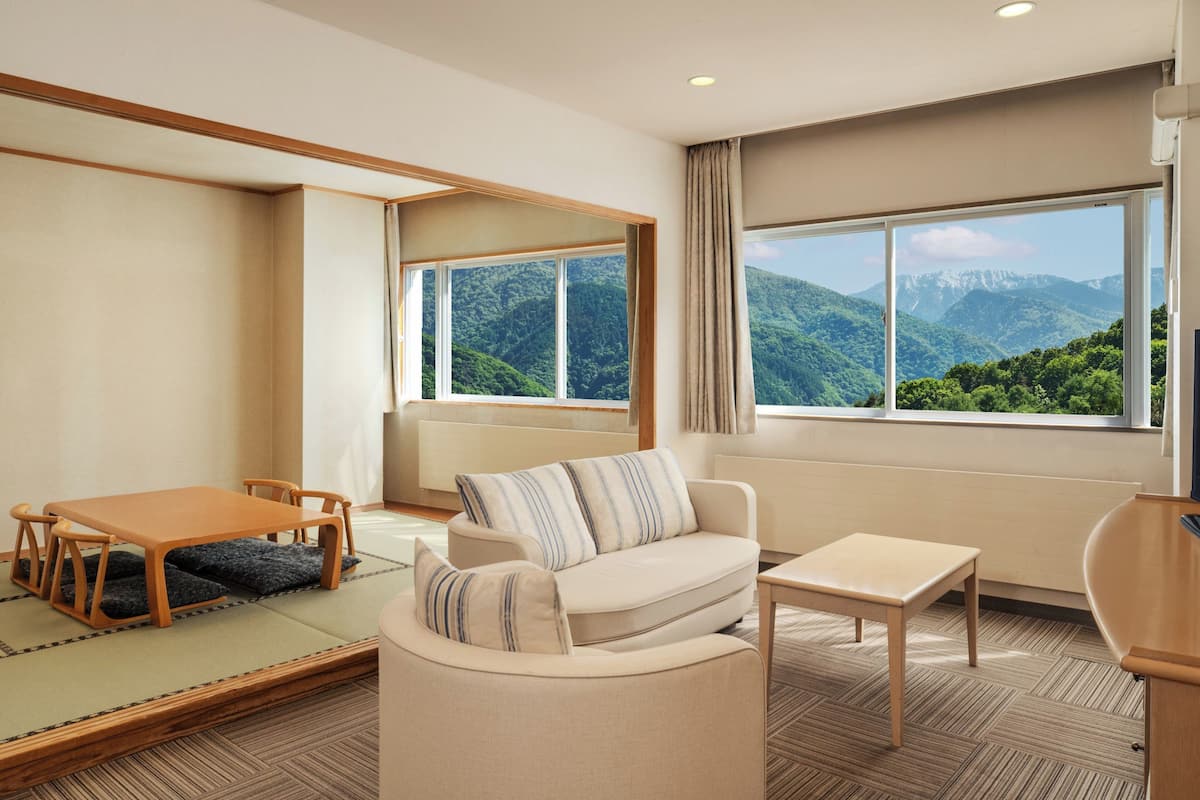 A modern hotel room with a sofa, a small table, a wooden chair, and a view of mountains through large windows.