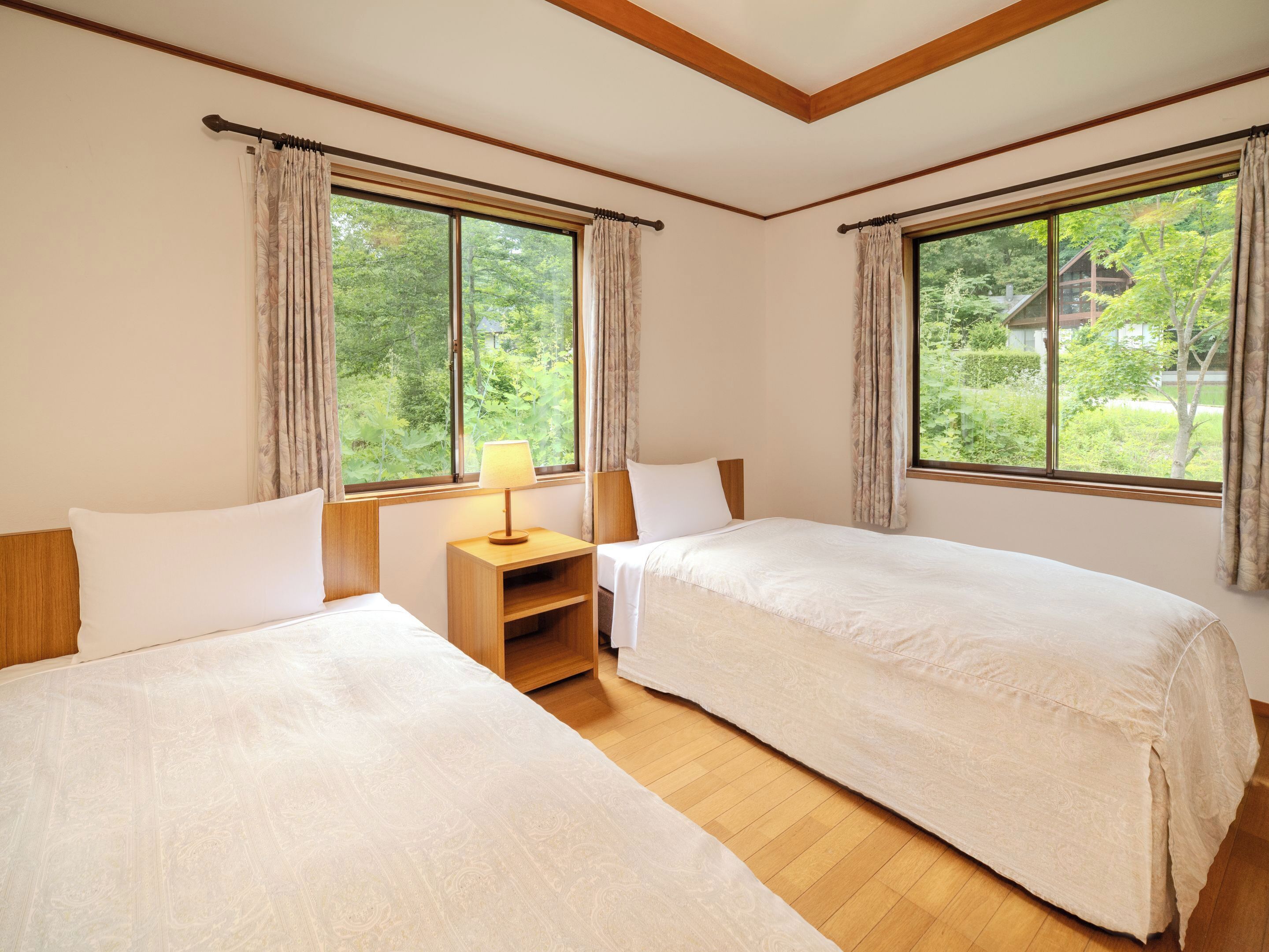 A room with two beds, a nightstand with a lamp, large windows with curtains, and a view of greenery outside.