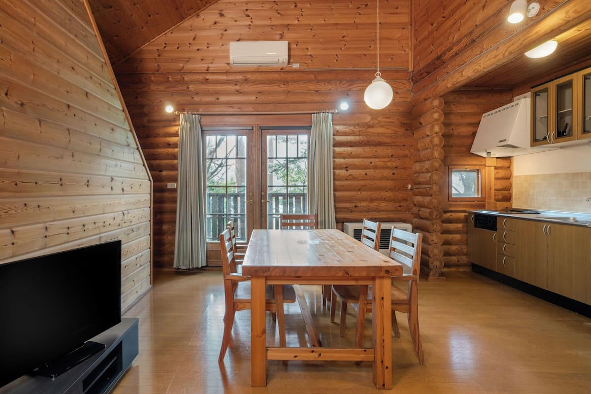 A log cabin interior with a dining table, chairs, a TV, and a kitchen area.
