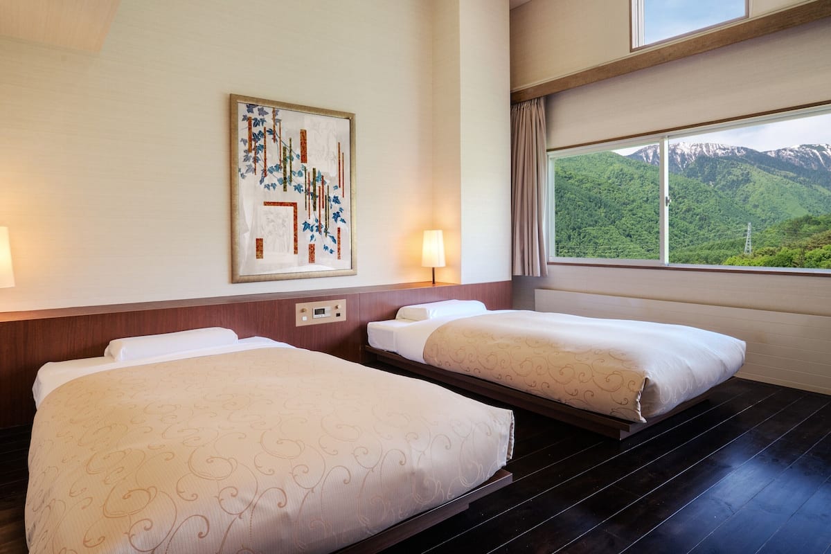 A hotel room with two beds, a painting on the wall, a window with a mountain view, and a control panel on the wall.