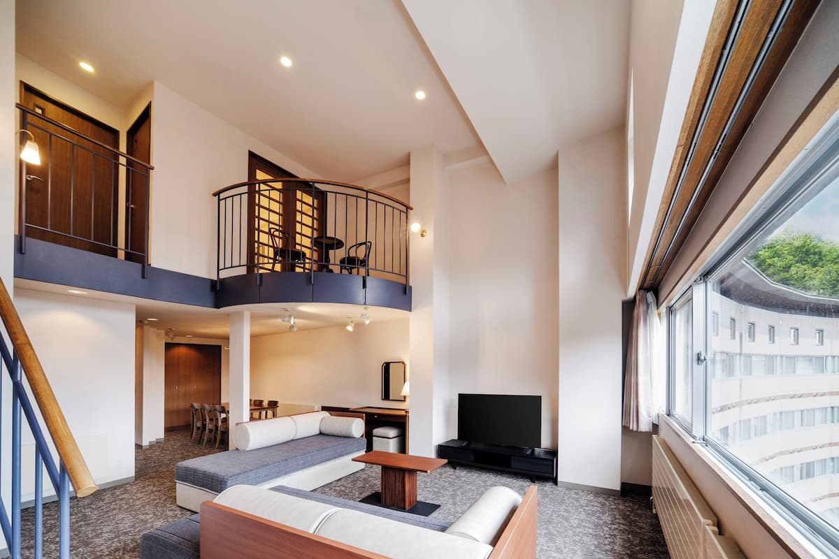A modern hotel room with a loft area, a sofa set, a dining table, and a TV.