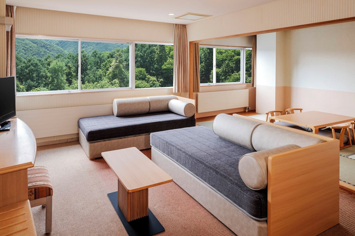 A modern room with a sofa, a small table, a TV, and large windows offering a forest view.