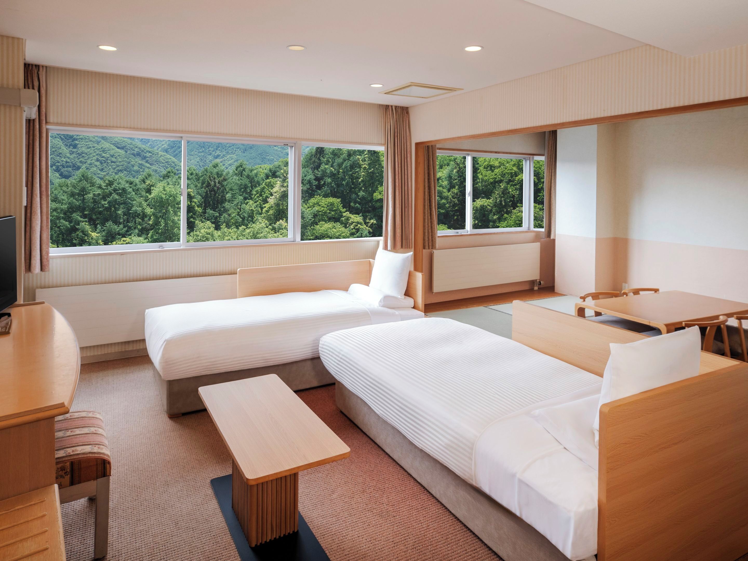 Une chambre d’hôtel avec deux lits, un bureau, une chaise et de grandes fenêtres donnant sur la forêt.
