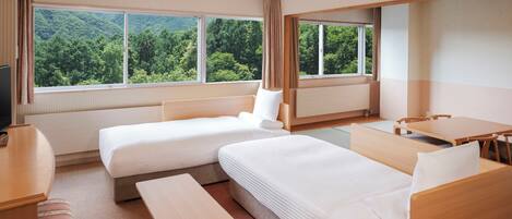 A hotel room with two beds, a desk, a chair, and large windows offering a forest view.