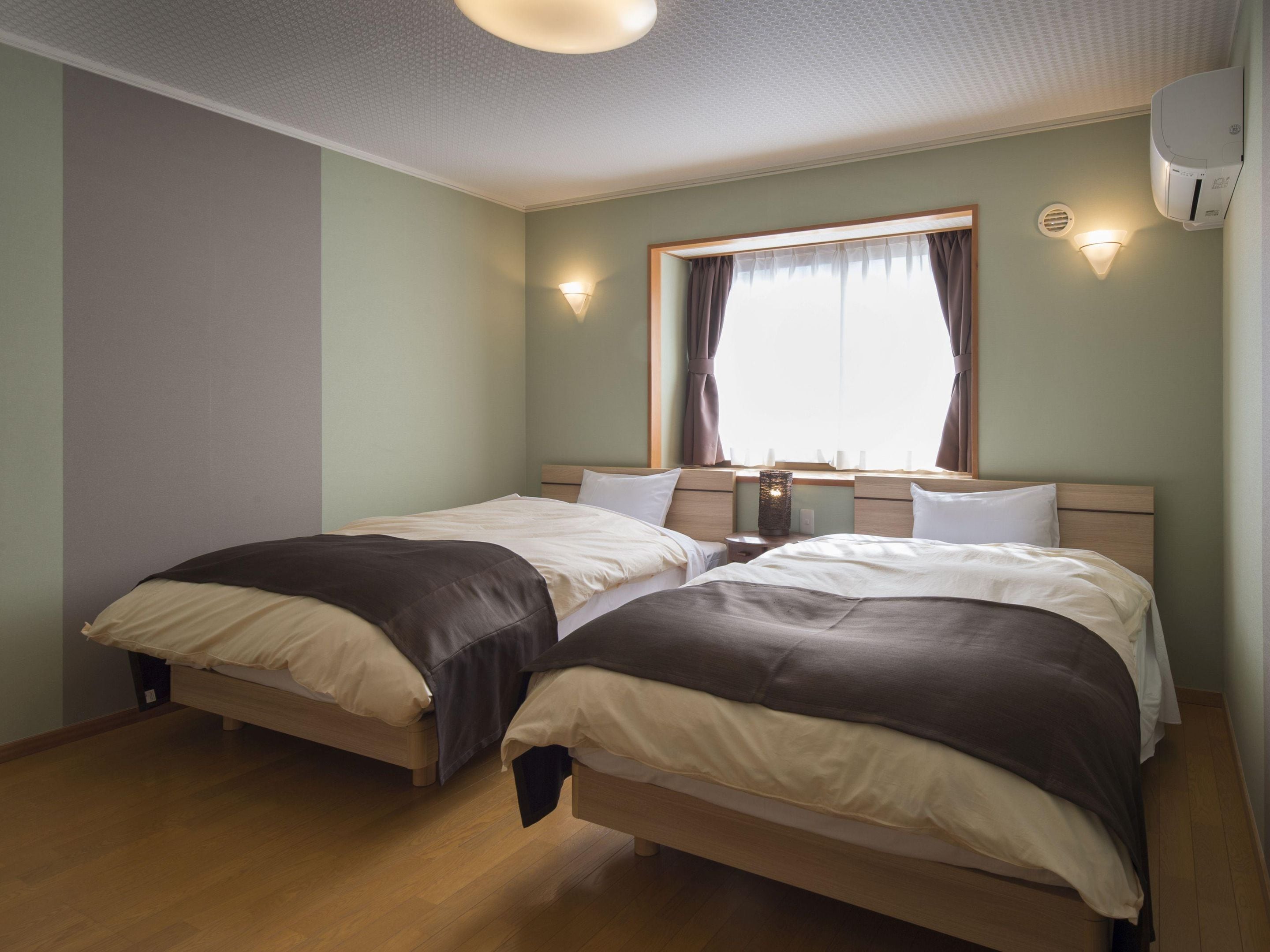 Une chambre double avec deux lits, une fenêtre avec des rideaux, un climatiseur fixé au mur et une table de chevet avec une lampe.