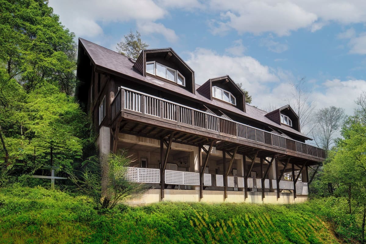 A two-story wooden house with a balcony, surrounded by lush greenery.