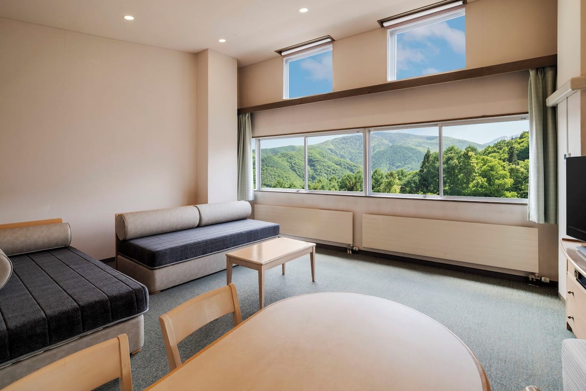 A room with a sofa, a table, and a view of a mountainous landscape through large windows.