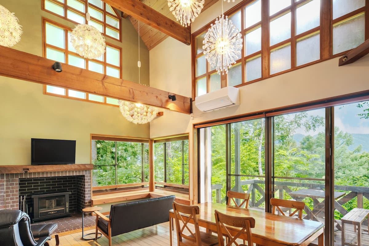 A spacious living area with a fireplace, wooden furniture, large windows, and a balcony with a view of the outdoors.