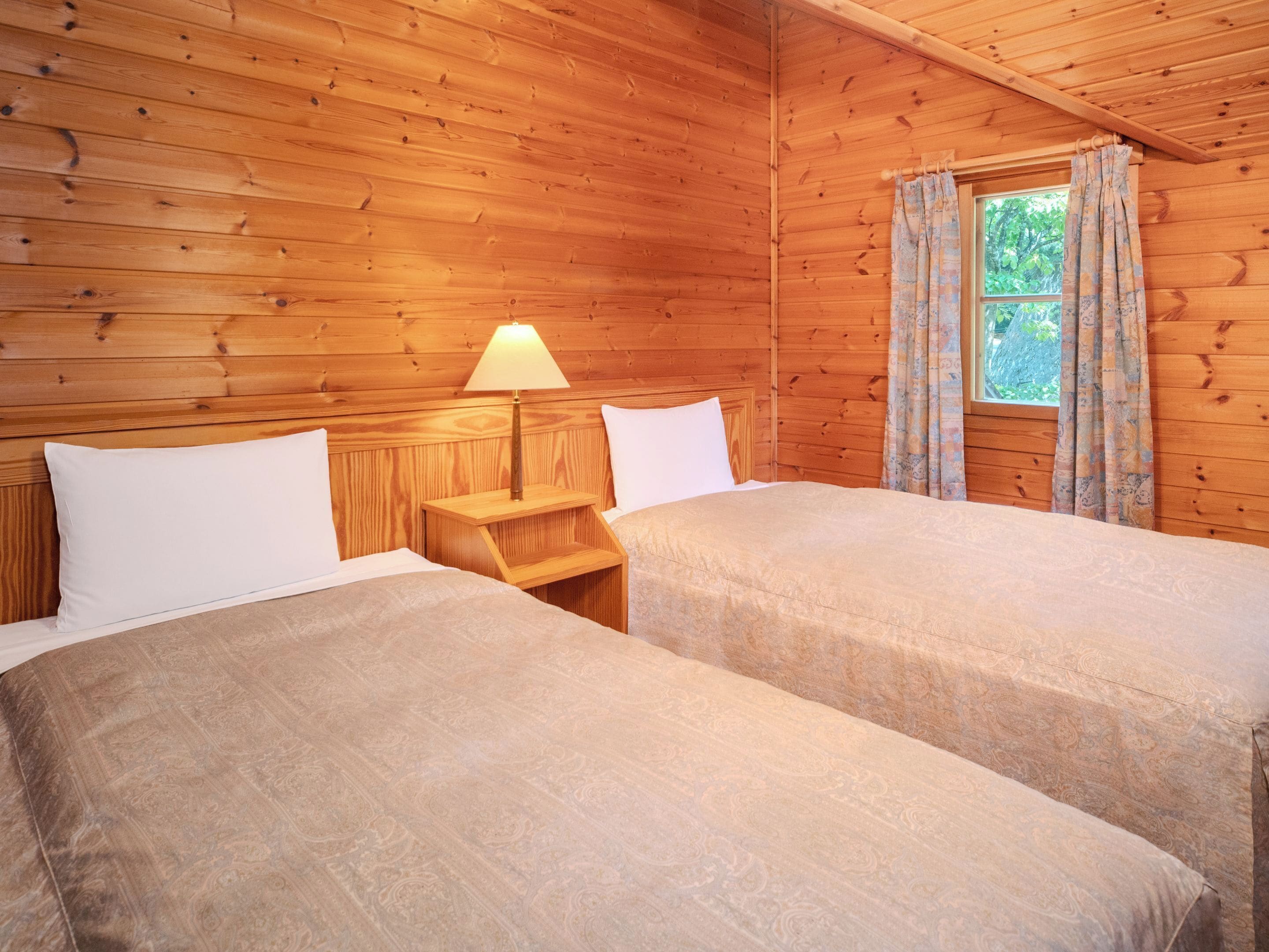 A wooden cabin room with two beds, a nightstand with a lamp, and a window with curtains.