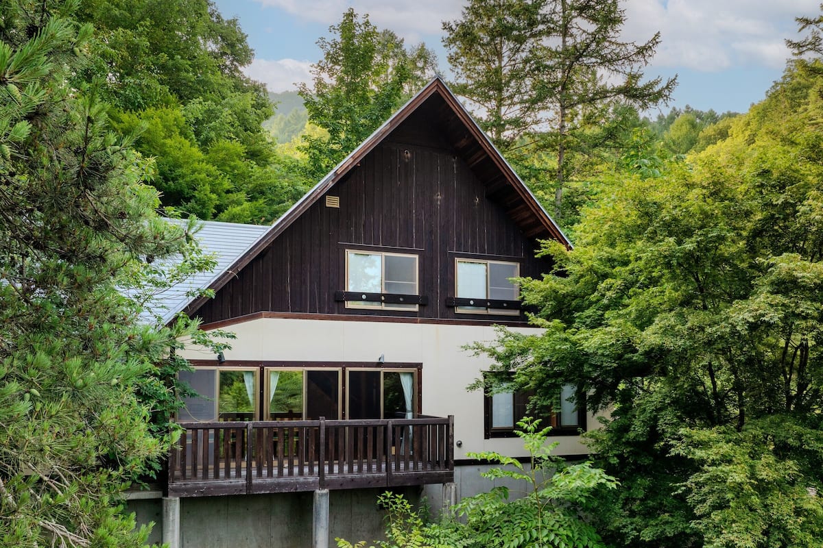 A two-story wooden cabin with a balcony, surrounded by dense forest.