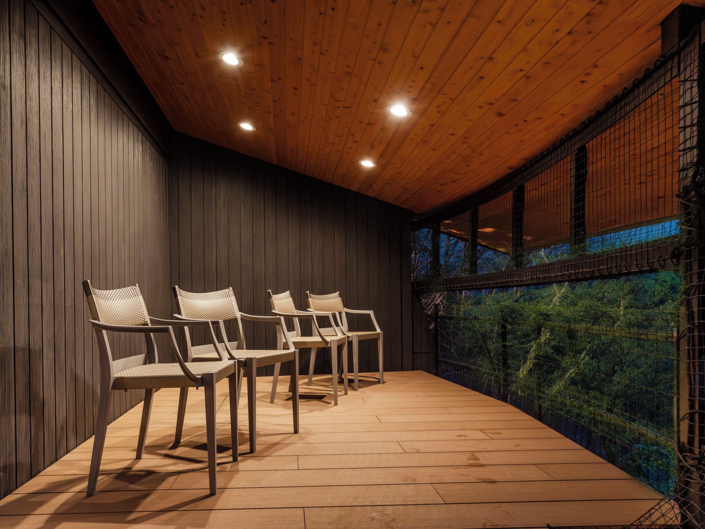 A room with wooden flooring, four wicker chairs, and a large window with a view of a forest.