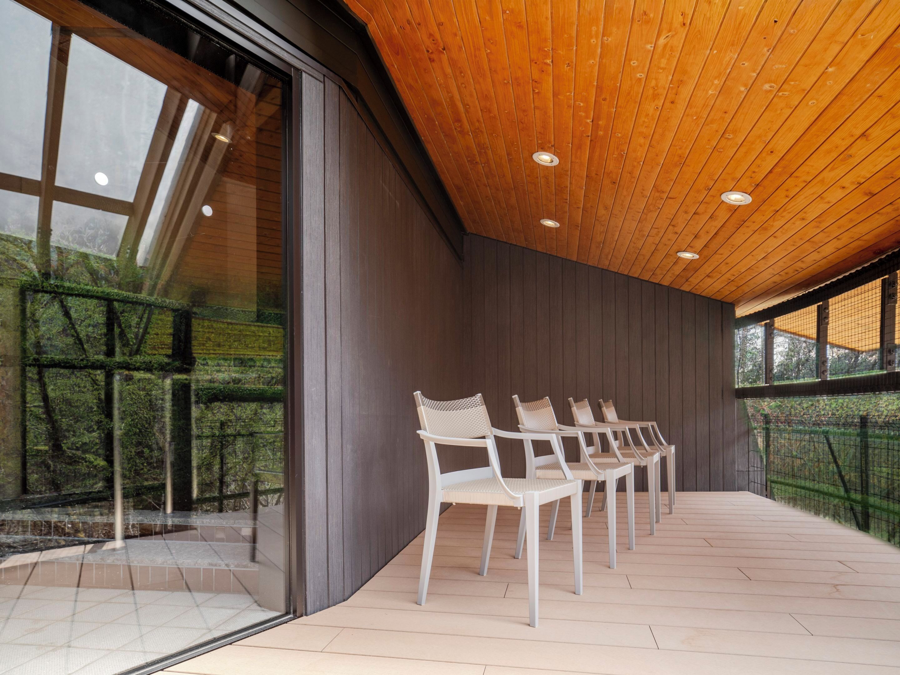 Ein modernes Interieur mit Holzbalkendecke, großen Fenstern und einer Reihe weißer Stühle.